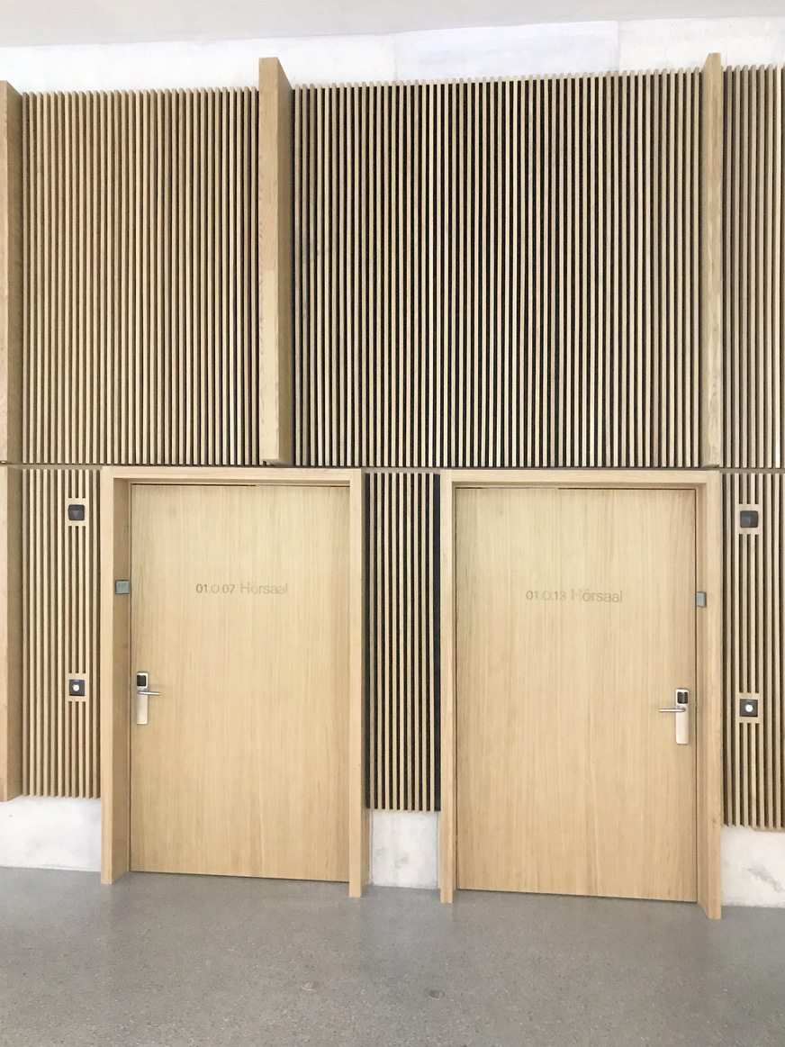 Die Flurwände der Hörsäle im ersten und zweiten Obergeschoss sind mit Lamellen aus Eichenholz ausgeführt. In die vertikale Struktur sind auch die Öffnungen der Überströmelemente eingebunden.