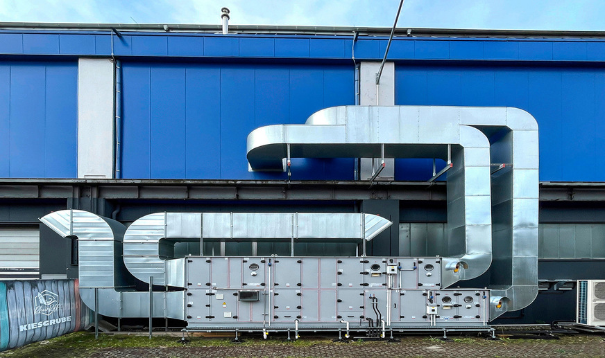 Das RLT-Gerät KG Top mit geschwungenen Zu- und Fortluft-führenden Kanälen steht im Außenbereich auf dem Boden neben der Halle - es hätte auf dem geschwungenen Hallendach nicht platziert werden können.